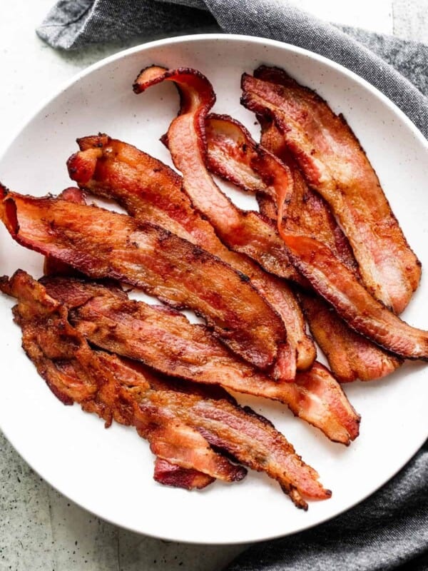 strips of air fryer bacon arranged on a white plate, with a gray tea towel placed underneath the plate.