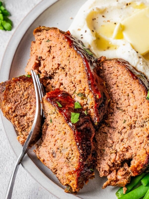 A fork taking a bite out of a slice of air fryer meatloaf on a plate with mashed potatoes and green beans.