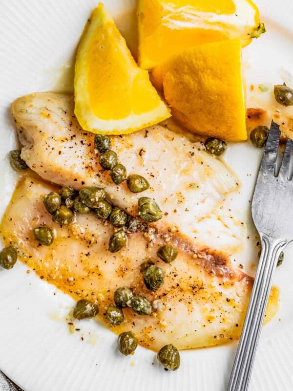 Air fryer tilapia on a plate with lemon caper sauce and fresh lemons.