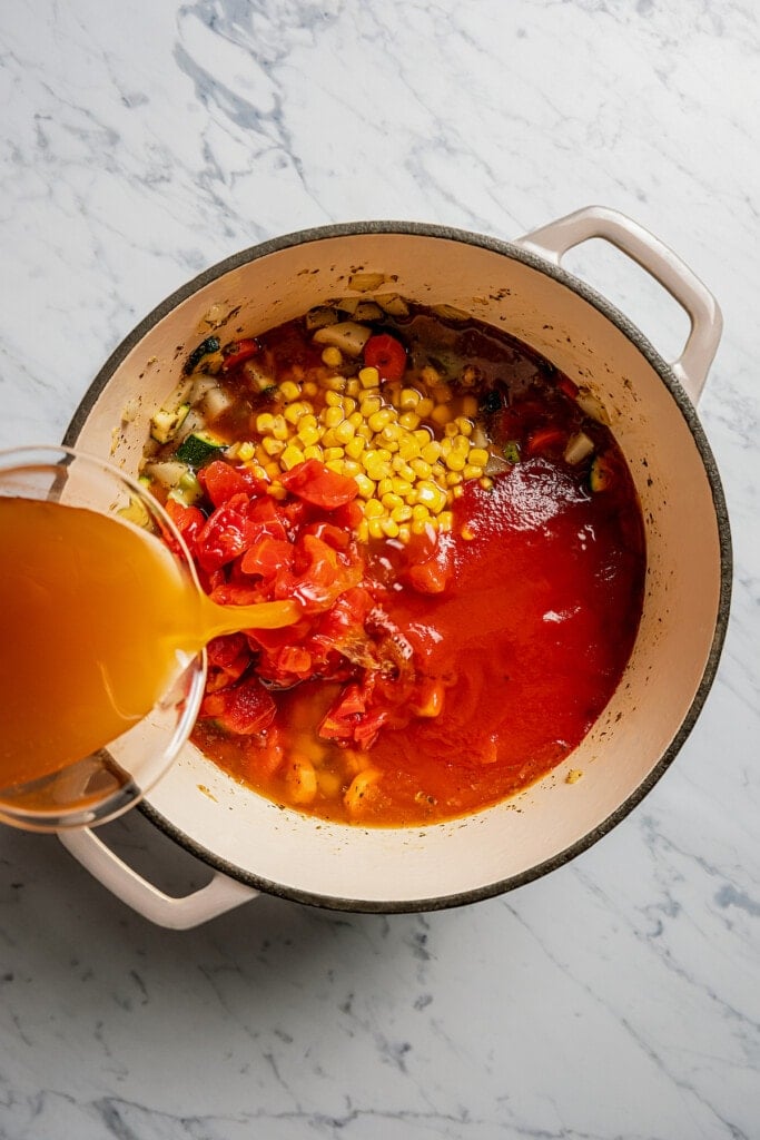 Adding corn, diced tomatoes, tomato sauce, and chicken broth to sauteed veggies to make soup.