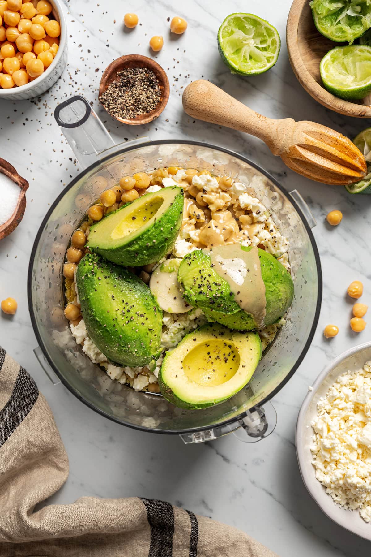 Avocado hummus ingredients combined in a blender, surrounded by garnishes, crackers, small bowls of seasonings, and a bowl of feta.