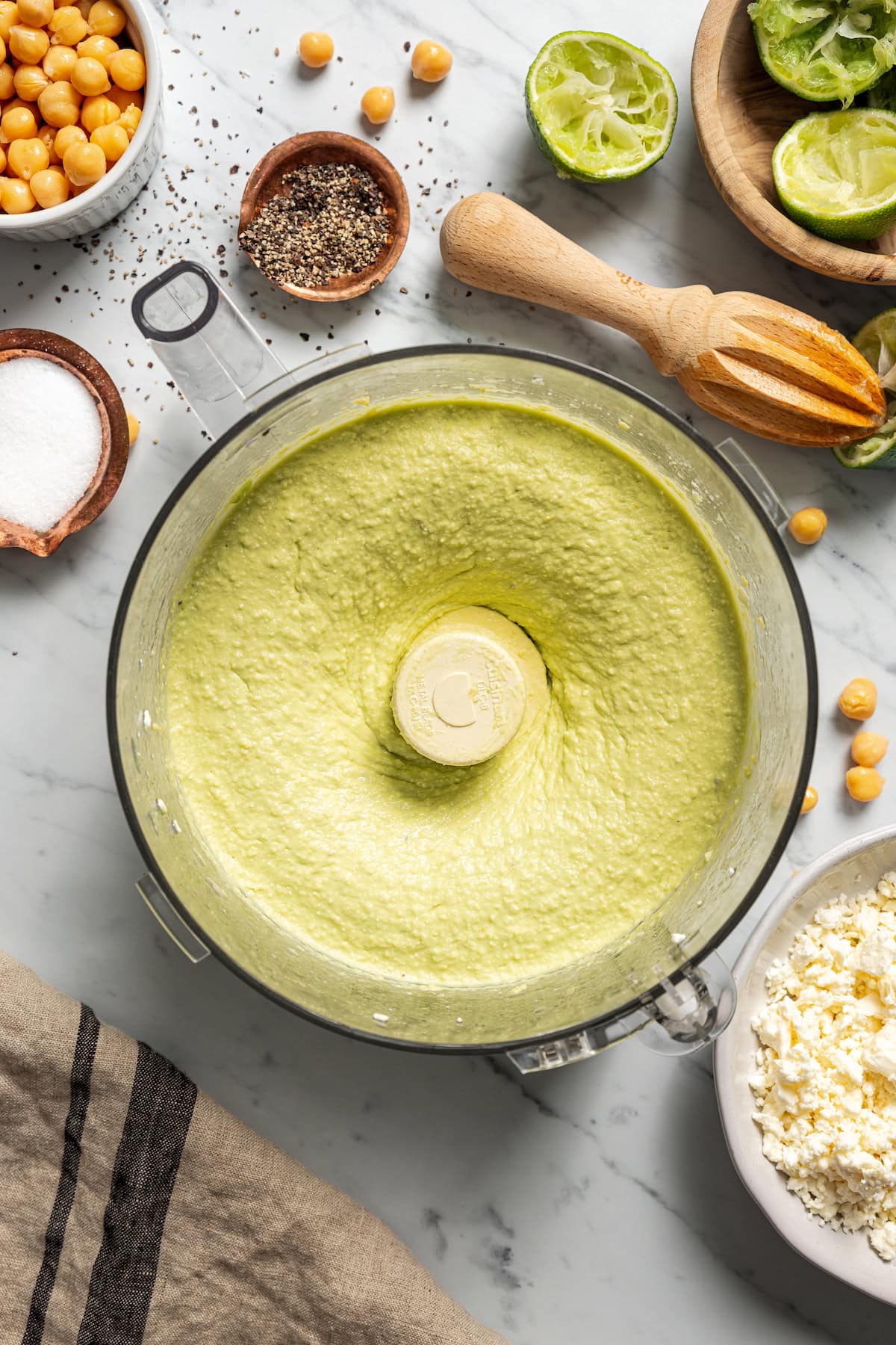 Blended avocado hummus in a blender, surrounded by garnishes, crackers, small bowls of seasonings, and a bowl of feta.