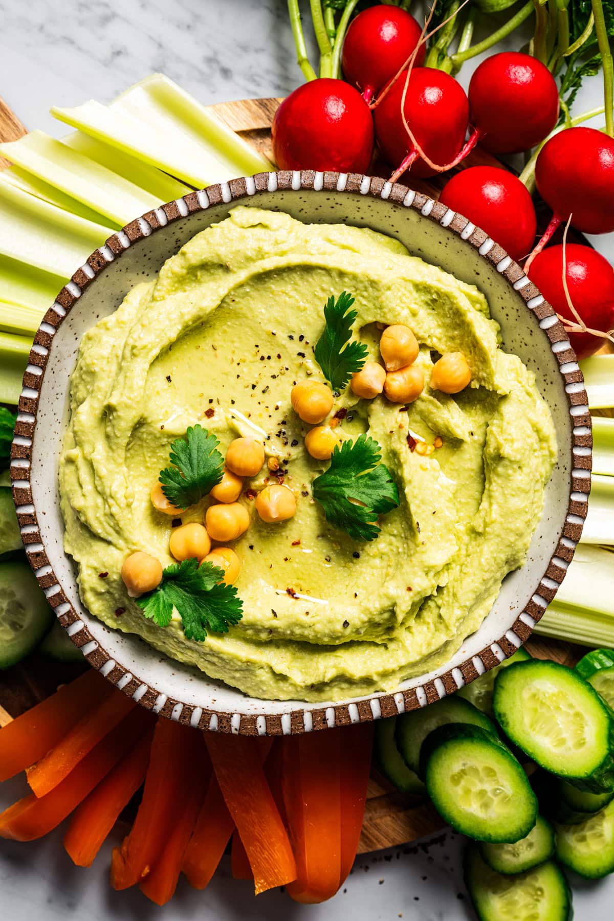 A bowl of hummus dip garnished with chickpeas, cucumber slices, and parsley, surrounded by crudites.