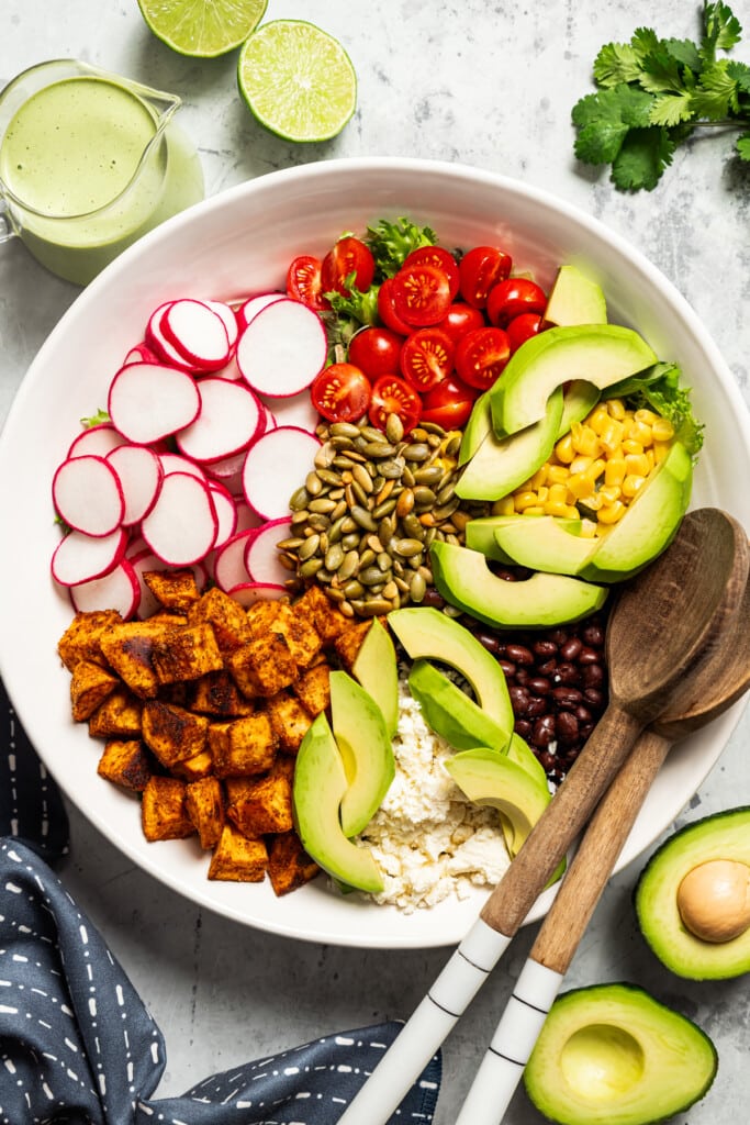 Adding veggies, black beans, pepitas, avocado, and queso fresco to a bowl full of salad greens.