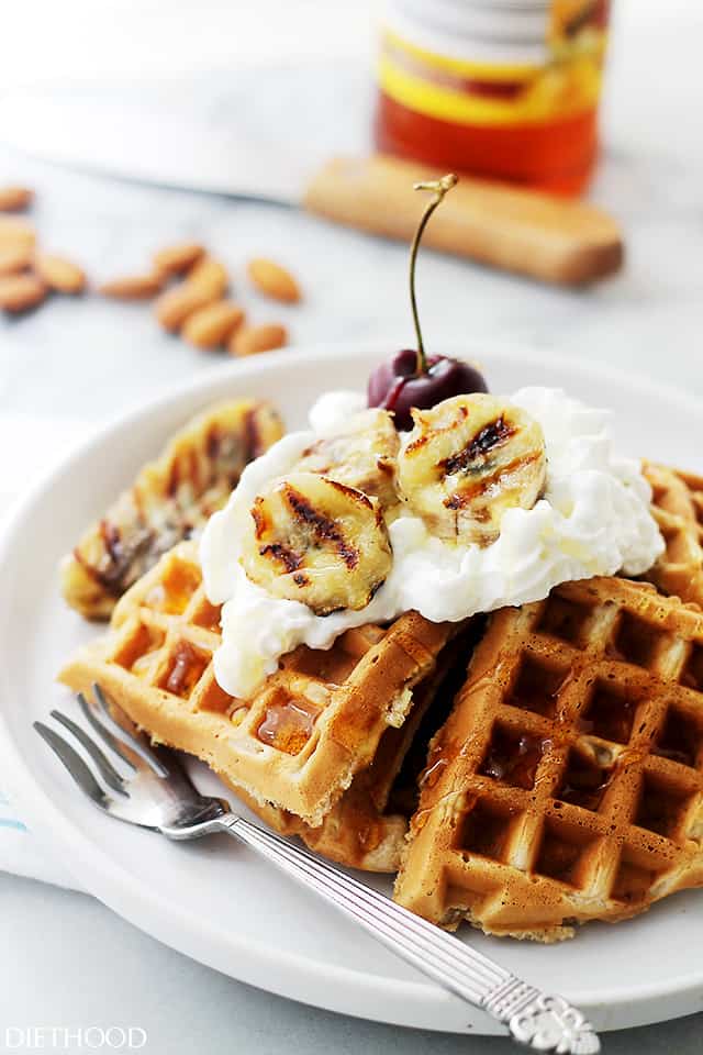 A stack of banana bread waffles topped with whipped cream and grilled banana slices on a white plate next to a fork.