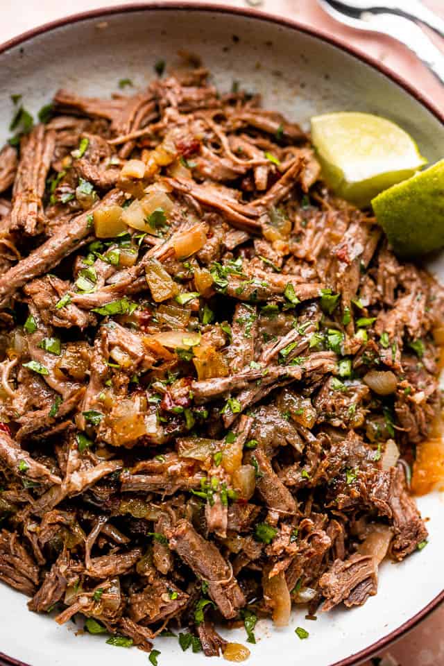 Close up of pulled beef barbacoa served in a large bowl with lime wedges.