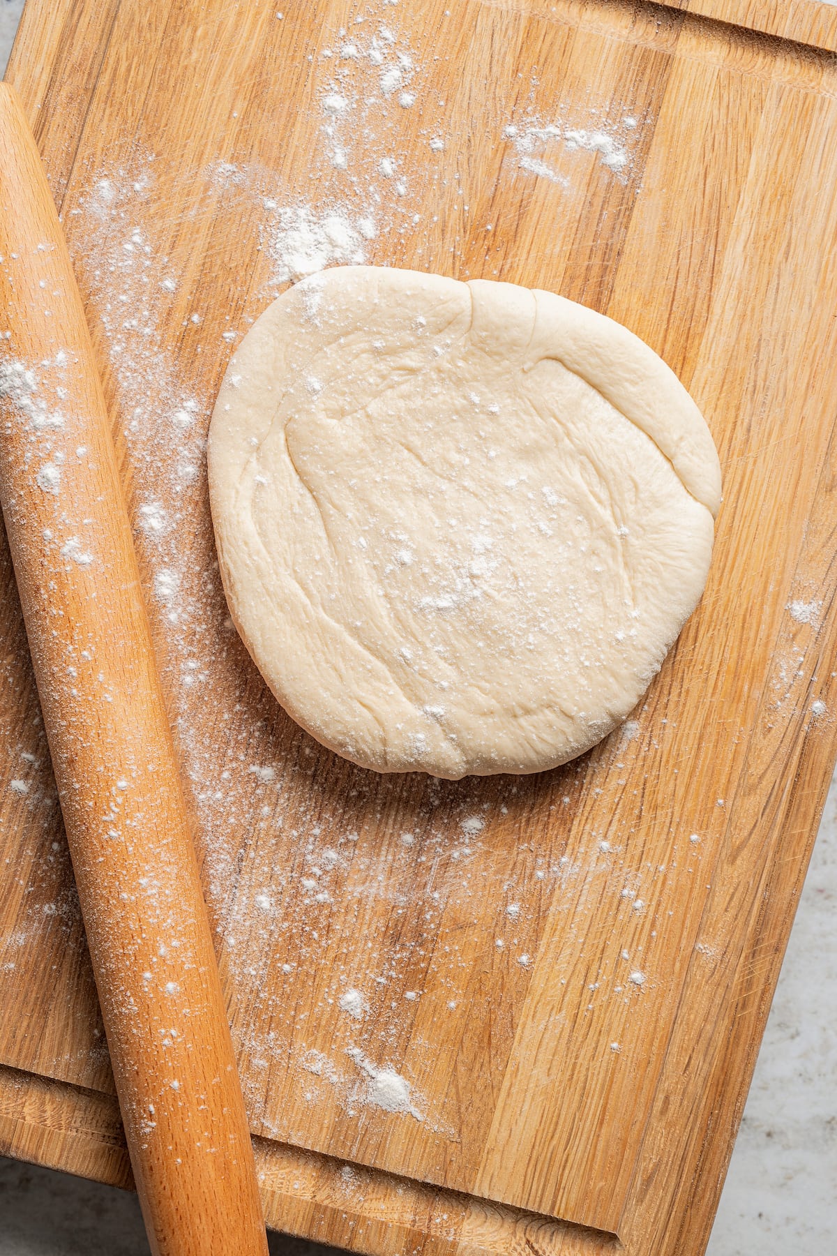 Pizza dough turned out on a floured wooden cutting board next to a wooden rolling pin.