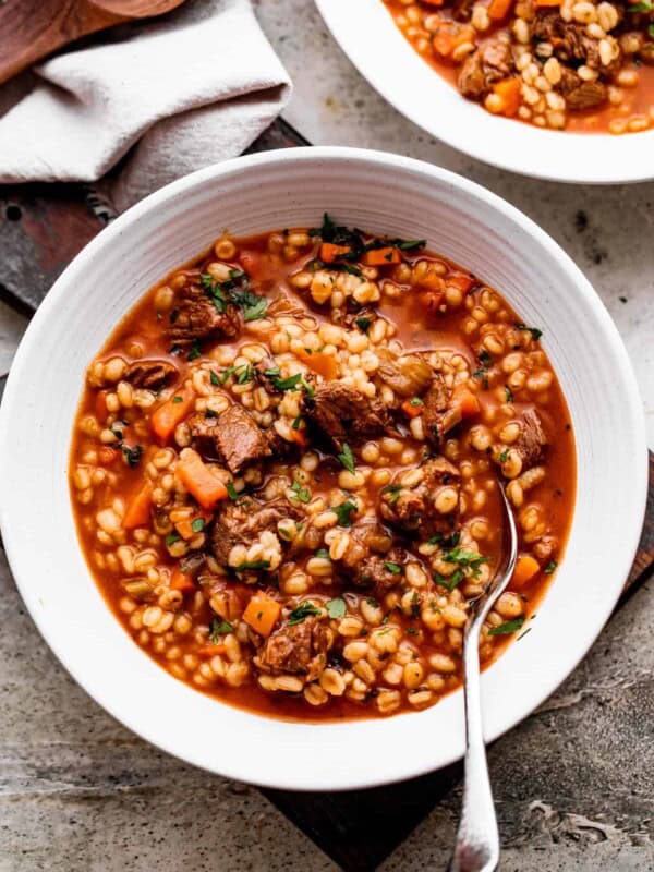 side shot of two bowls filled with beef barley soup and a spoon dipped in the soup.