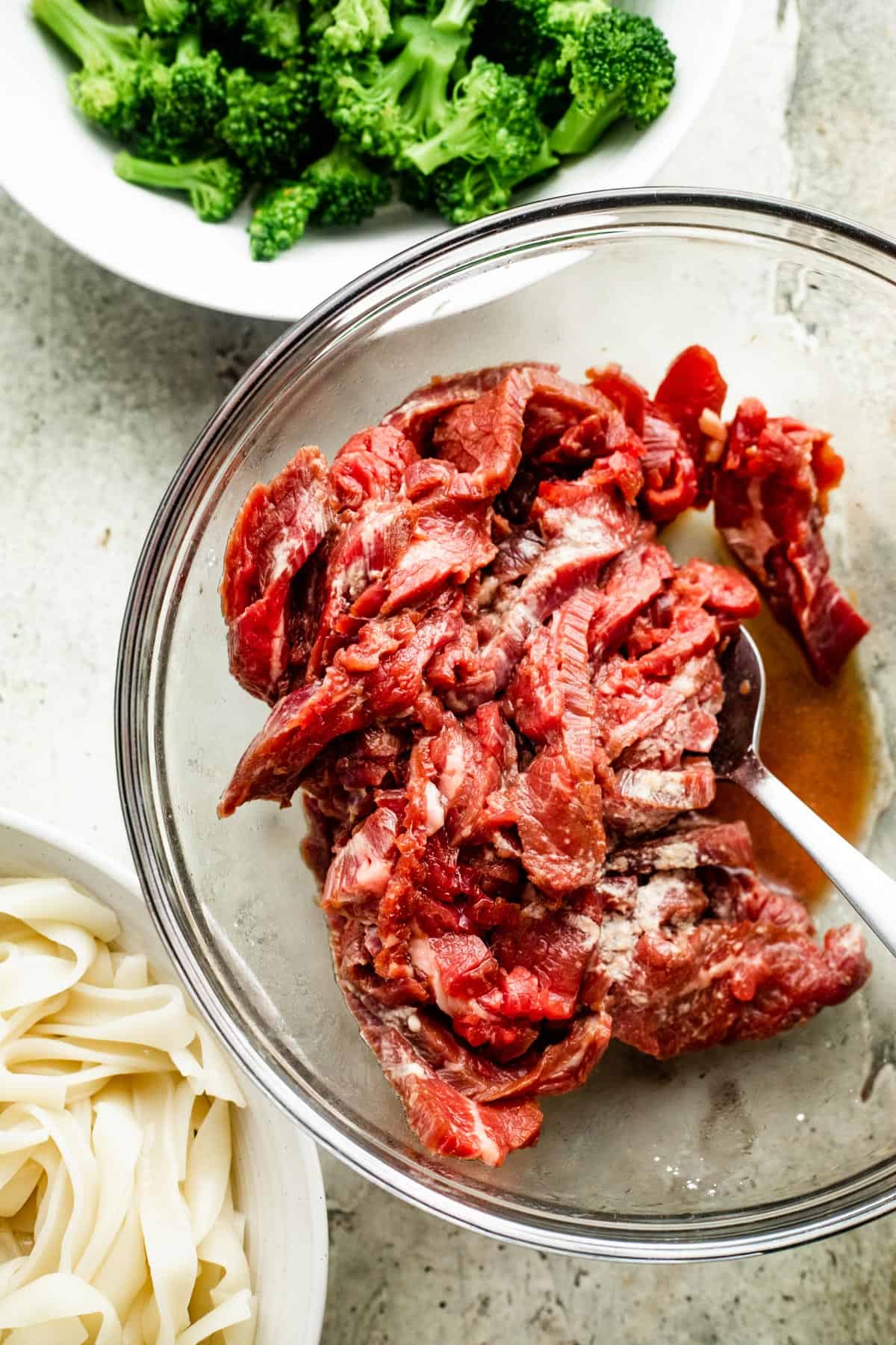 overhead shot of a glass bowl with sliced flank steak, a white bowl above the glass bowl filled with broccoli florets, and a white bowl below the glass bowl filled with wide rice noodles.