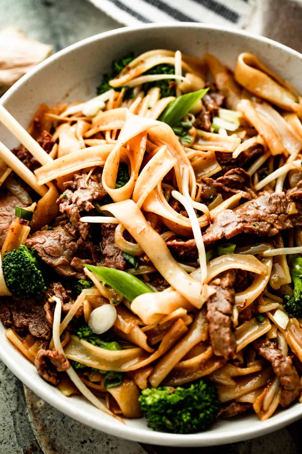 up close overhead shot of a white bowl with beef chow fun and two chopsticks resting on the left side of the bowl.