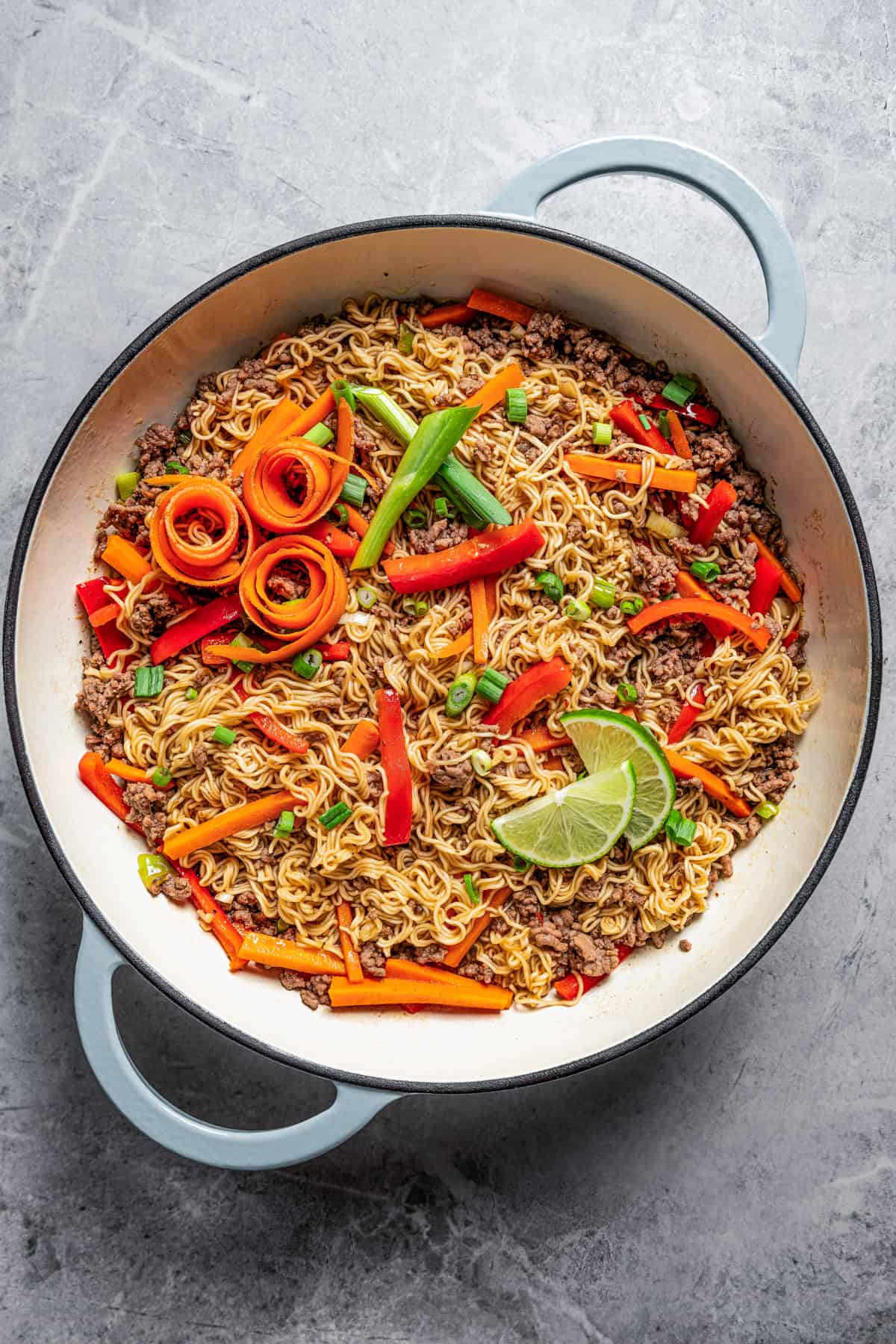 Beef ramen noodles in a large pot garnished with carrot ribbons, limes, and scallions.