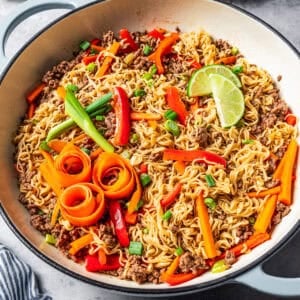 Beef ramen noodles in a large pot garnished with carrot ribbons, limes, and scallions.