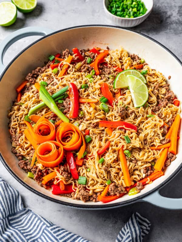 Beef ramen noodles in a large pot garnished with carrot ribbons, limes, and scallions.