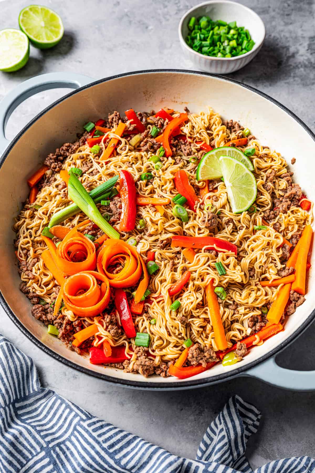 Beef ramen noodles in a large pot garnished with carrot ribbons, limes, and scallions.