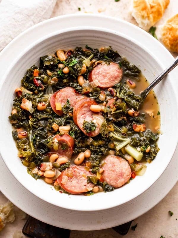 overhead shot of a white bowl filled with black eyed peas, kale, sliced andouille sausage, and broth. Bread slices are placed near the top right of the soup bowl.