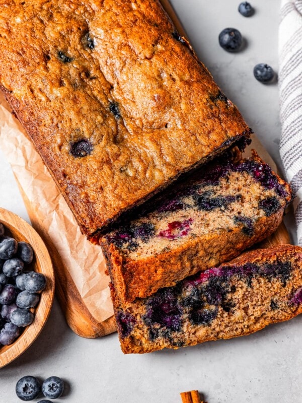 Overhead view of a loaf of blueberry banana bread cut into slices next to a bowl of blueberries.