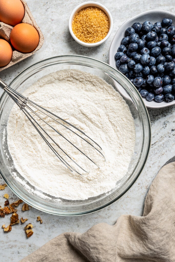 Overhead image of prep bowls: one bowl is filled with flour, another with raw sugar, and a third with fresh blueberries. Eggs are placed nearby.