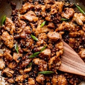 wooden spoon stirring through bourbon chicken while cooking in a stainless steel skillet.