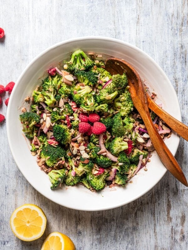 Overhead image of broccoli salad in a salad bowl with serving spoons and fork and a few fresh raspberries for garnish.