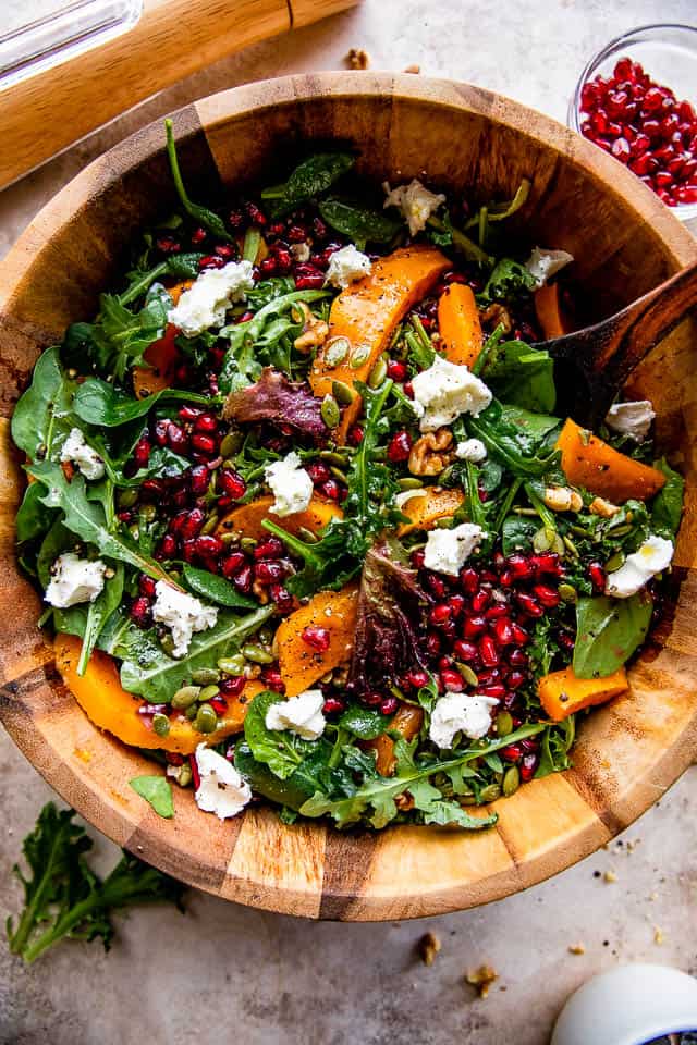 wide overhead shot of wooden salad bowl with salad greens, pomegranates, squash, and goat cheese