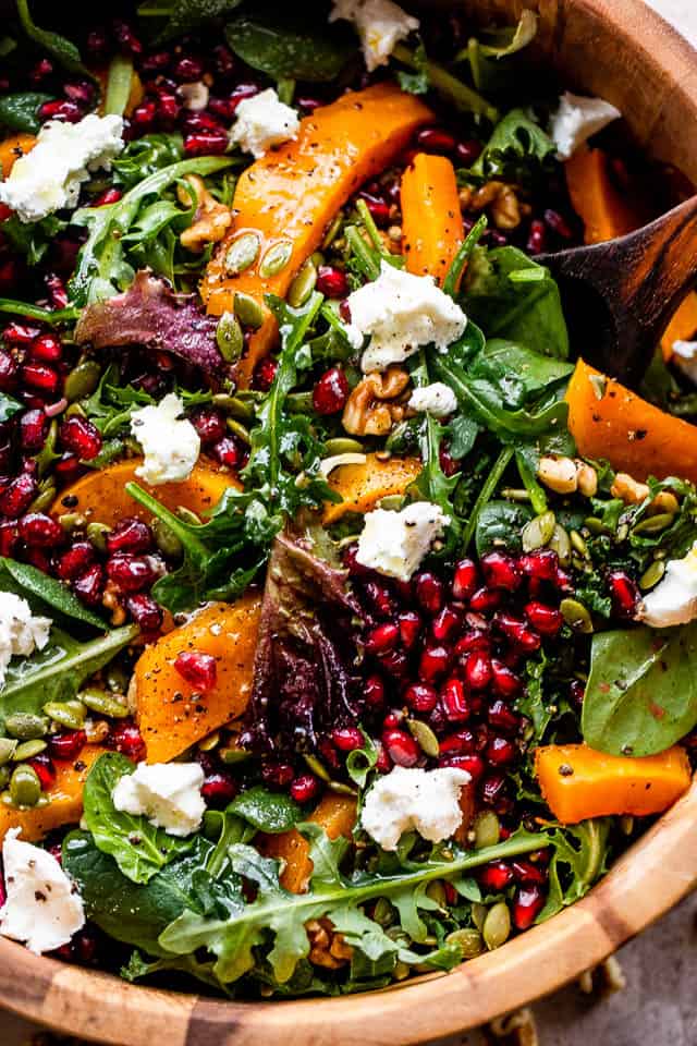 overhead shot of salad greens in a wooden salad bowl, and topped with squash, pomegranate arils, nuts, and goat cheese