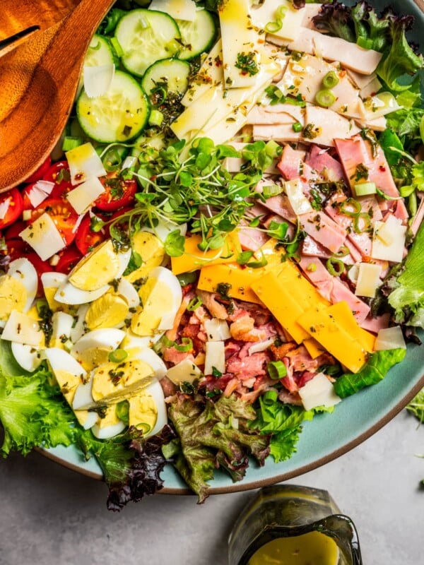 Chef salad in a large salad bowl dressed in Italian dressing next to wooden salad tongs.