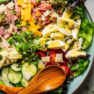 Chef salad in a large salad bowl dressed in Italian dressing, next to wooden salad tongs.