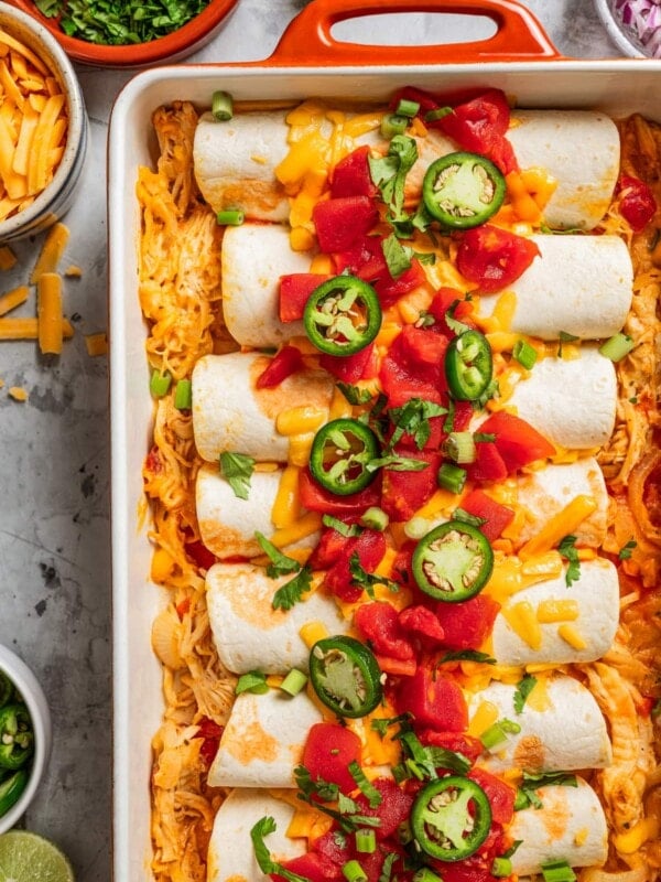 Enchiladas in a baking dish and topped with tomatoes, cheese, and sliced jalapenos.