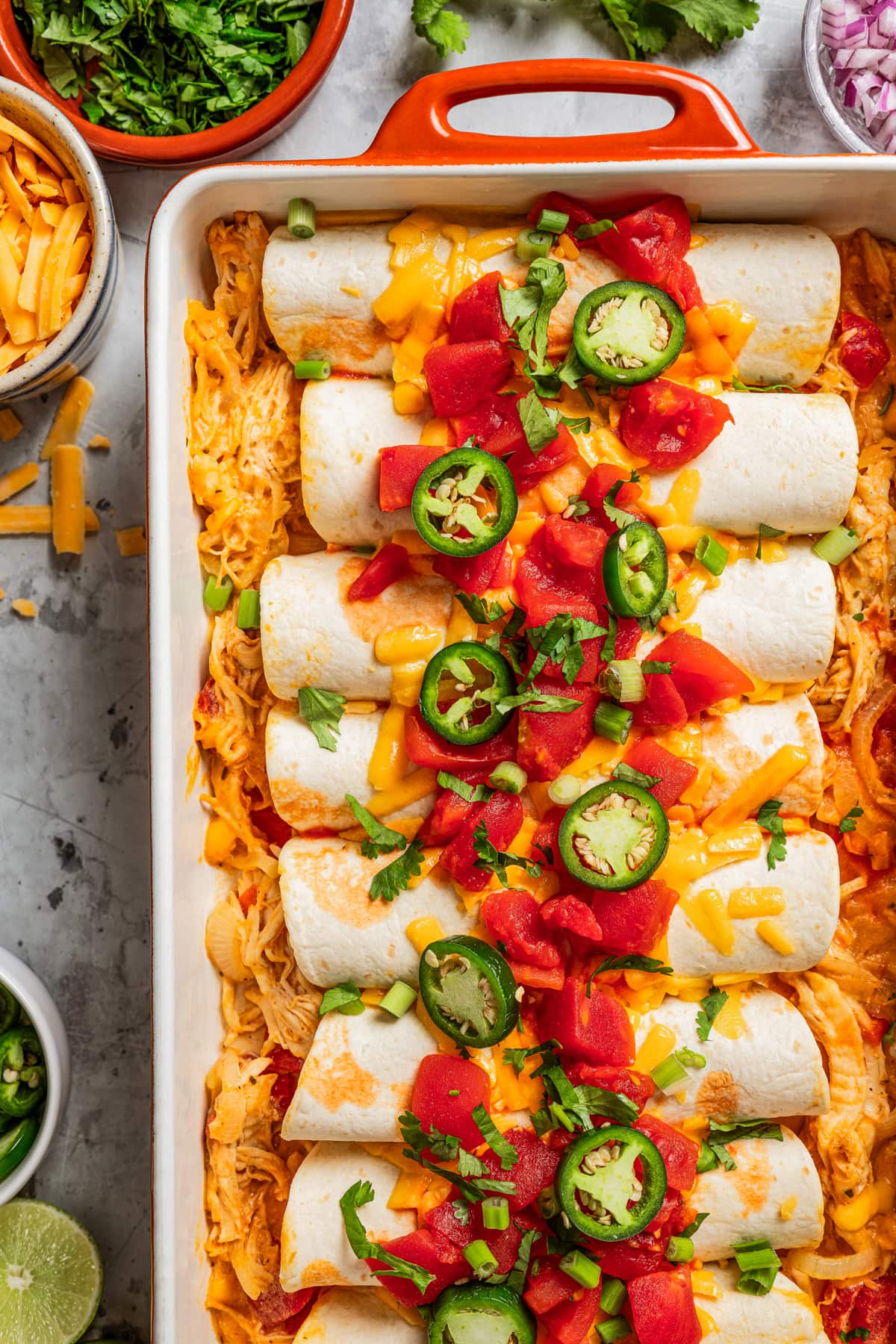 Enchiladas in a baking dish and topped with tomatoes, cheese, and sliced jalapenos.