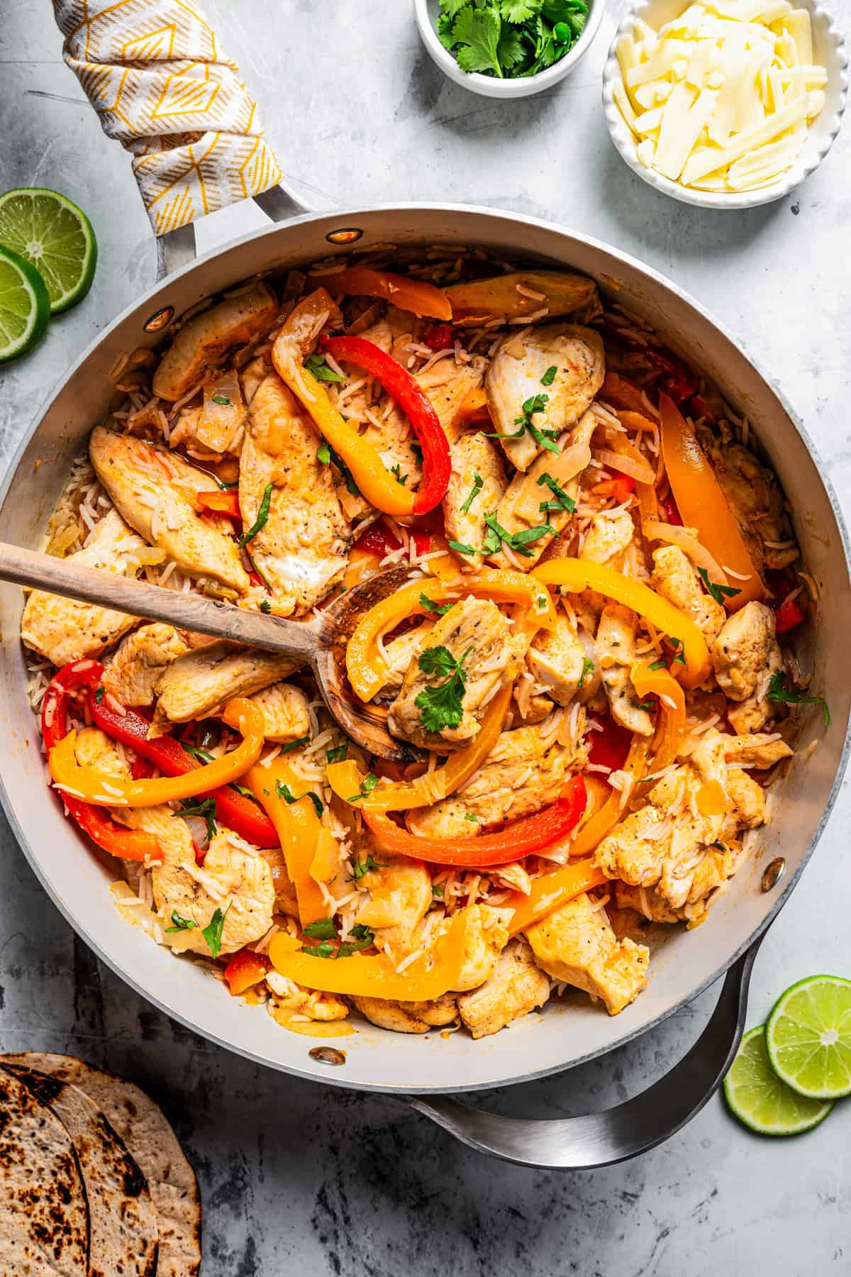 An overhead view shows a wooden spoon resting in a skillet of cooked sliced chicken, bell peppers, and onions.