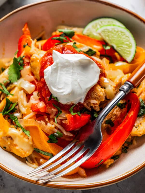 Close up of a chicken fajita bowl garnished with lime wedges and a dollop of sour cream, with a fork.