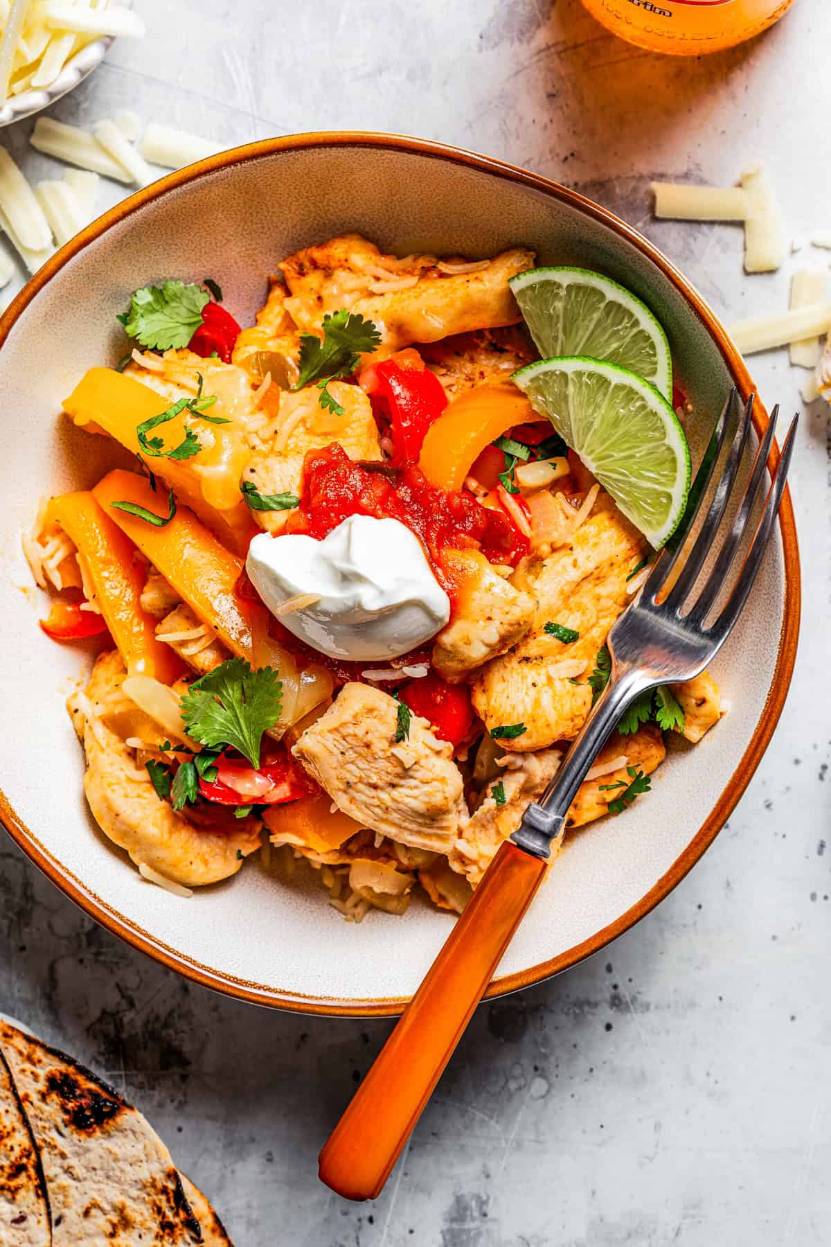 Photo of a bowl with chicken fajitas served over rice and garnished with a dollop of sour cream and lime wedges, with a fork, next to flour tortillas.