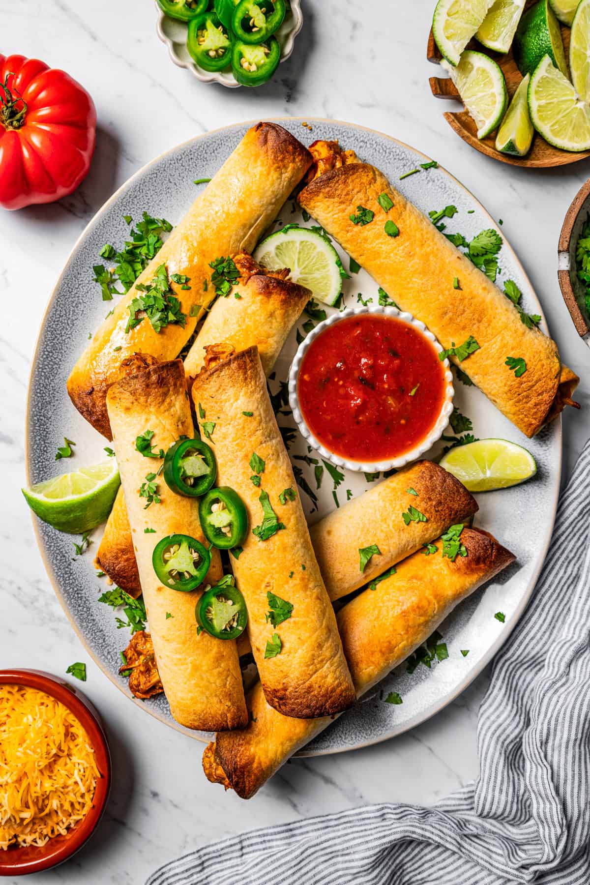 Flautas arranged on a serving platter with a bowl of dipping sauce. Slice jalapenos, lime wedges, tomatoes, and cheese are placed near the platter.