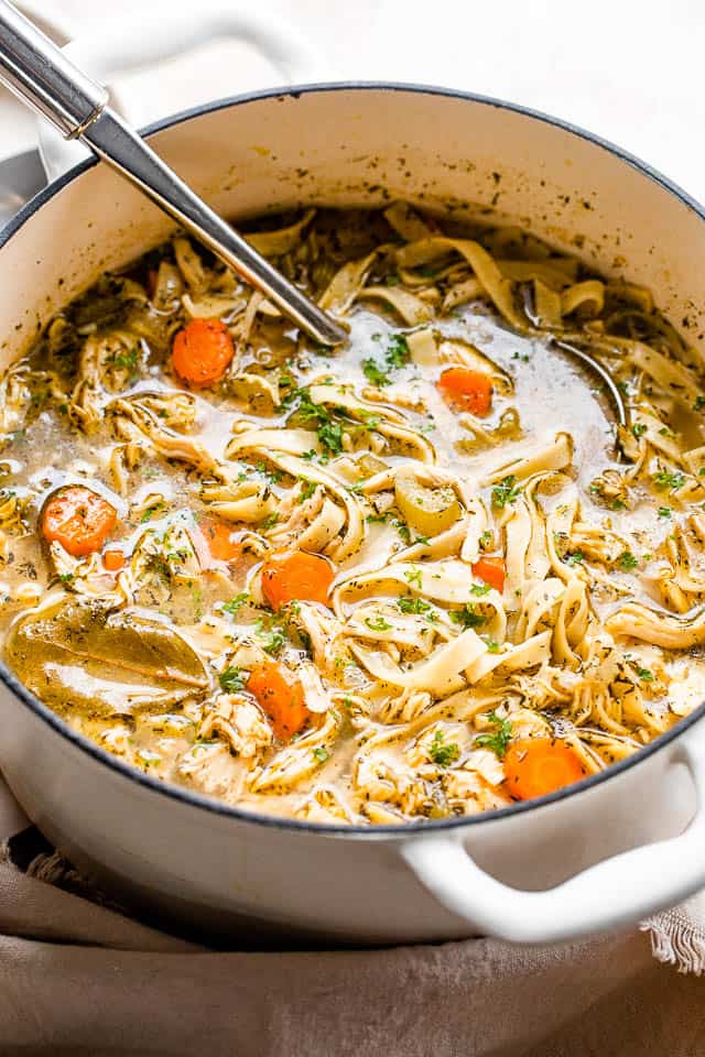 Side shot of chicken noodle soup in a white Dutch oven with a soup ladle inside the pot.