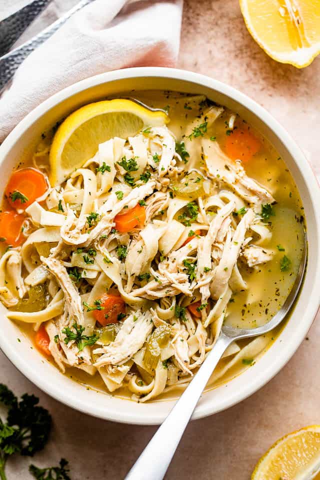 Overhead image of chicken noodle soup served in a soup bowl with a lemon wedge and a spoon inside the bowl.