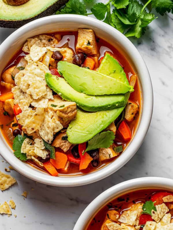 Overhead view of two bowls of tortilla soup topped with avocado and tortilla chips, next to a bowl of chips, a bowl of limes, half an avocado, and parsley.