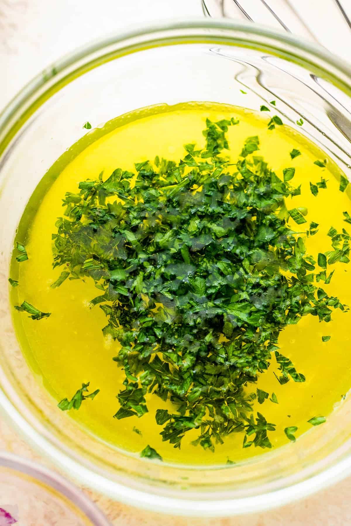 Olive oil, lemon juice, and chopped fresh herbs are prepped in a glass bowl for a salad dressing.