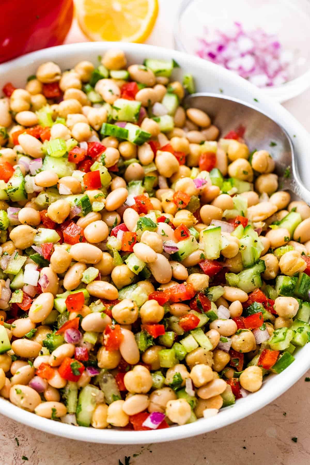 Salad bowl with garbanzo beans, diced cucumbers, celery, and red bell peppers. A spoon is resting inside the bowl.