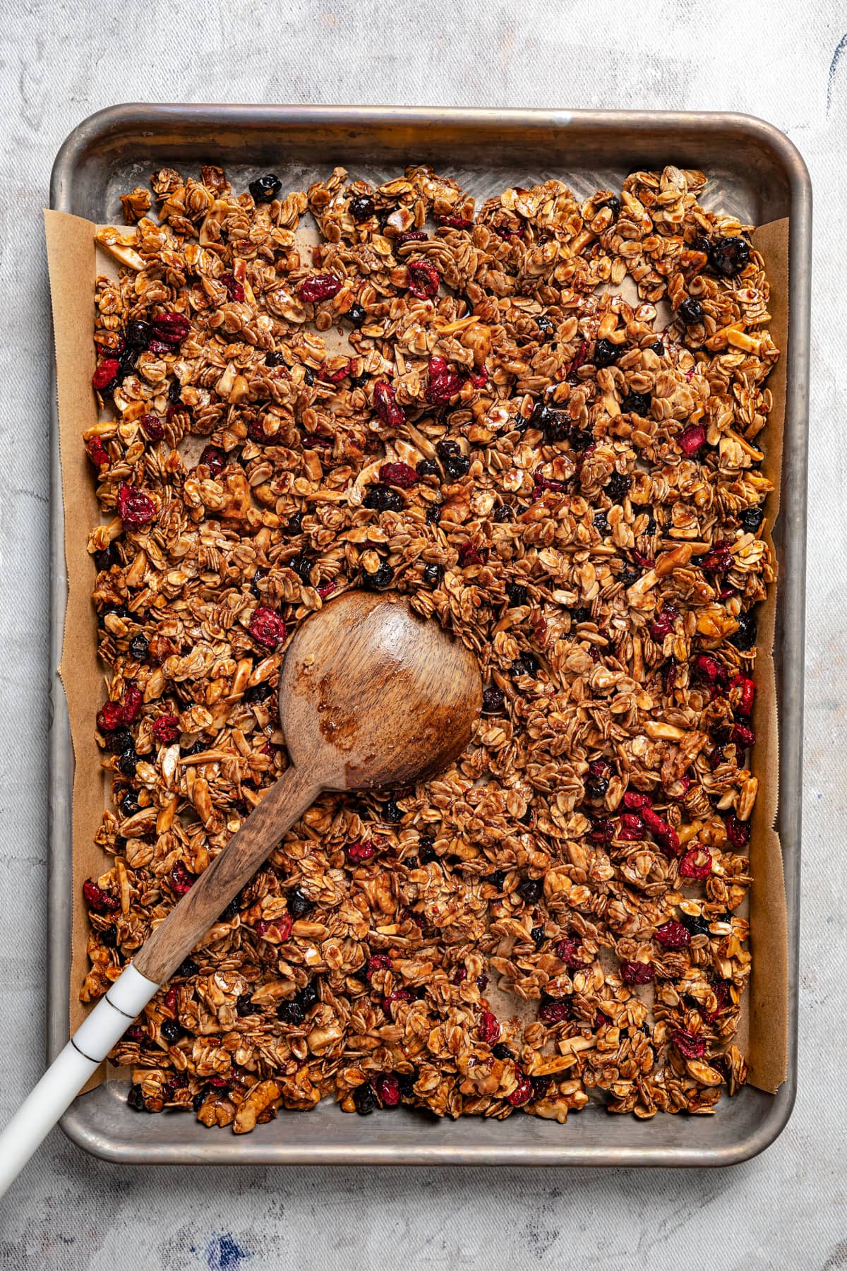 Overhead image of homemade granola on a baking sheet with a wooden spoon stirring through it.