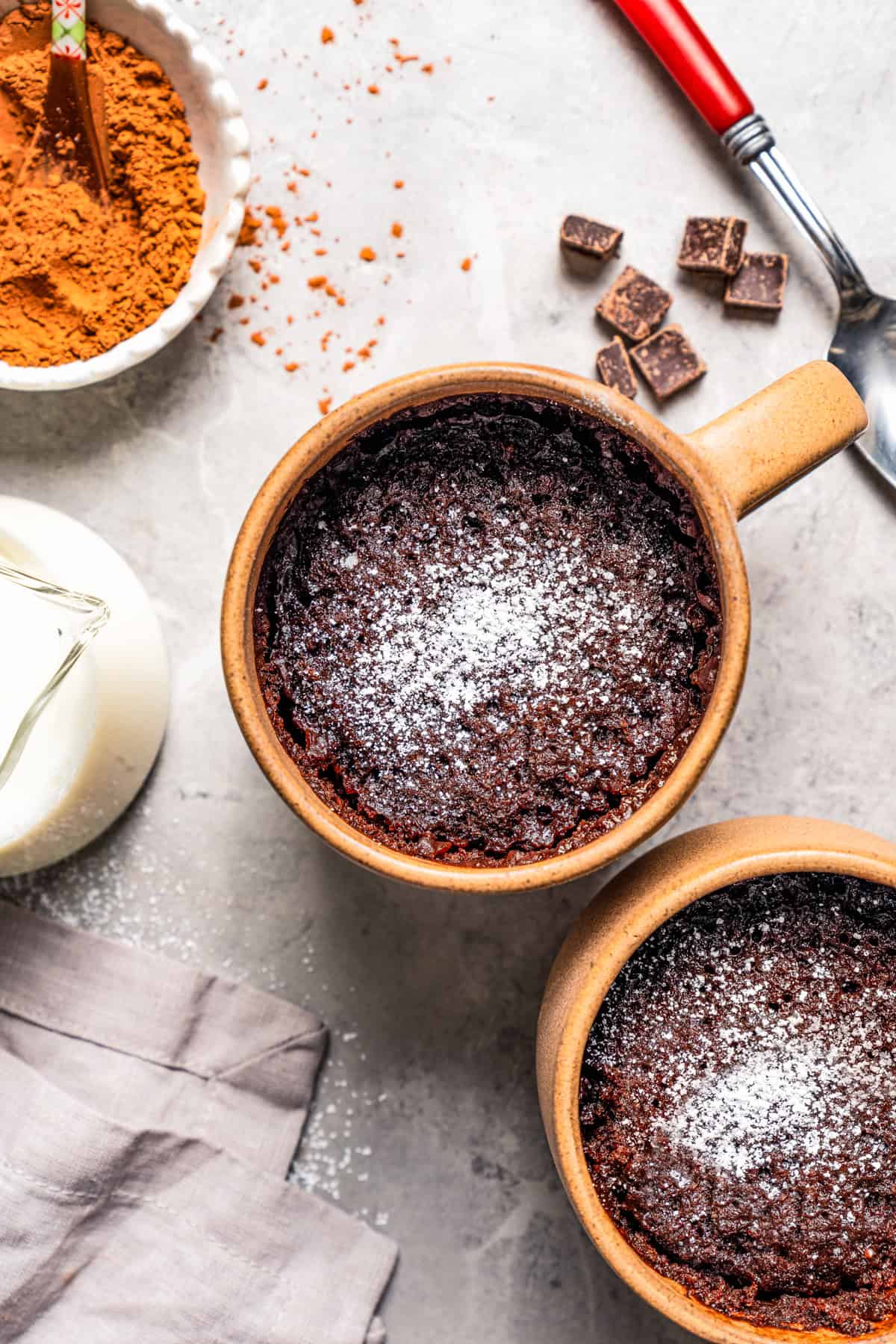 Overhead view of two chocolate mug cakes dusted with powdered sugar, next to a bowl of cocoa powder.