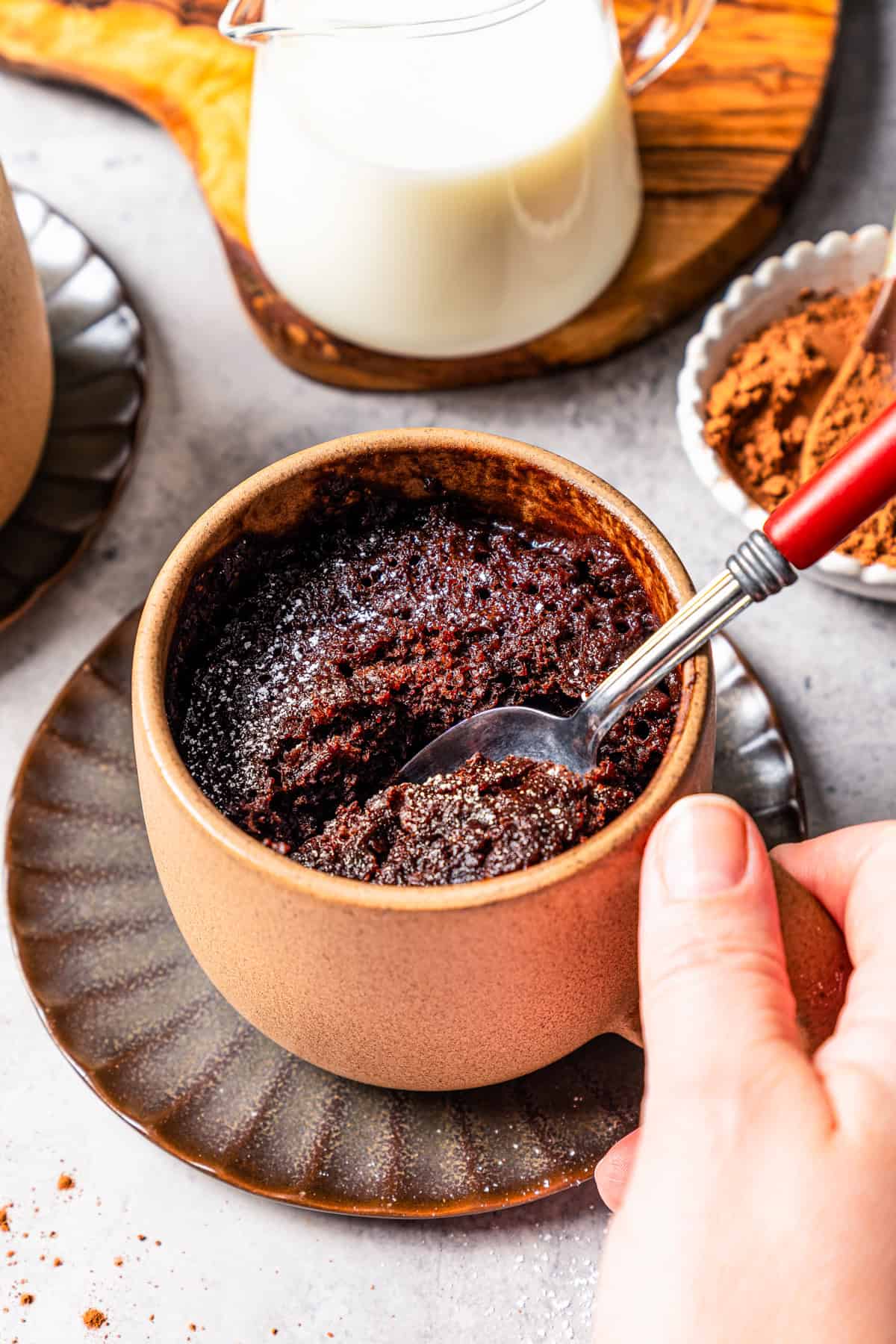 A hand holding the handle of a mug filled with chocolate cake, with a spoon.