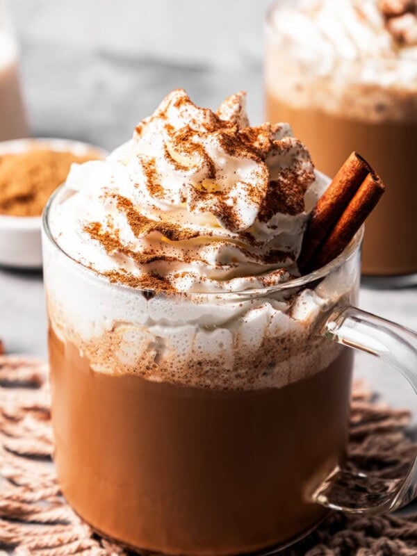 A cinnamon dolce latte in a glass mug garnished with whipped cream and a cinnamon stick, dusted with cinnamon, with a second latte in the background.