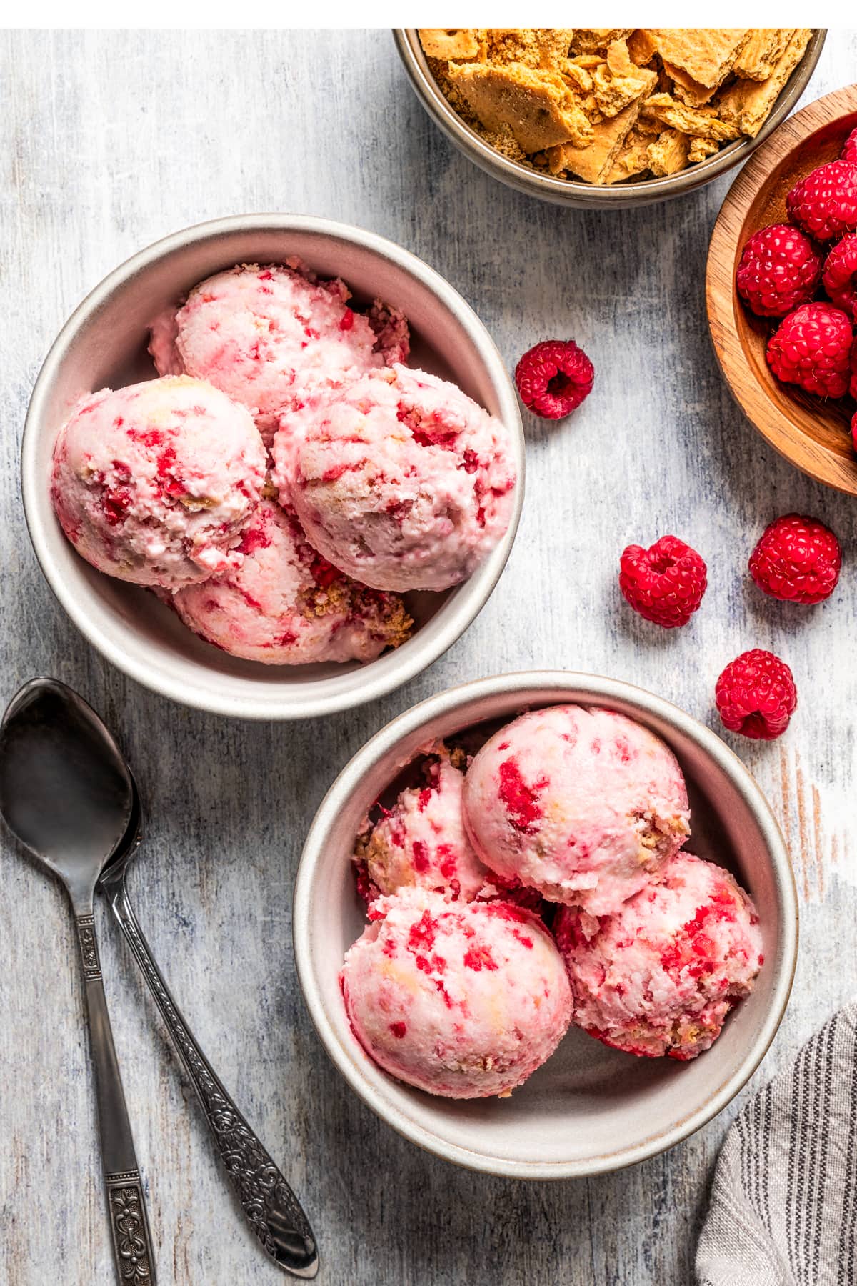 Two bowls of raspberry cottage cheese ice cream near a bowl of raspberries and a bowl of graham crackers.