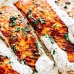 Fillets of salmon, pan-seared to a dark golden-brown, with a creamy sauce and chopped herb garnish.