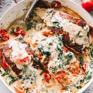 Overhead image of a skillet with three salmon fillets in a cream sauce mixed with baby spinach and cherry tomatoes.