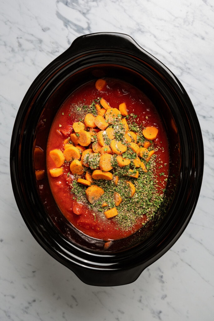 Loading a crockpot with carrots, canned tomato products, chicken stock, and seasonings.