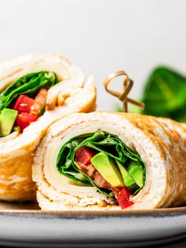 Closeup of a stuffed egg white wrap rolled up with cheese, turkey, avocado, salad, and tomatoes, and served on a plate.