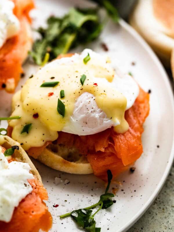 an english muffin topped with a slice of smoked salmon and a poached egg; top is garnished with hollandaise sauce and chopped chives.