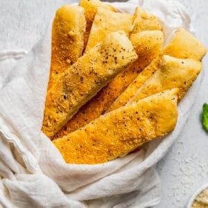 Garlic breadsticks wrapped with a white cloth, next to a bowl of garlic dipping sauce.