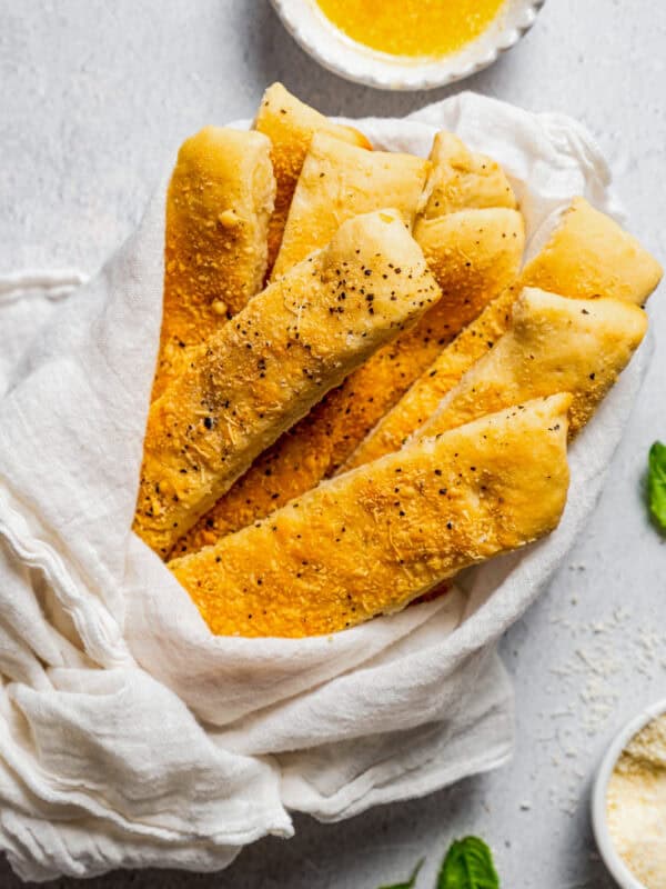 Garlic breadsticks wrapped with a white cloth, next to a bowl of garlic dipping sauce.